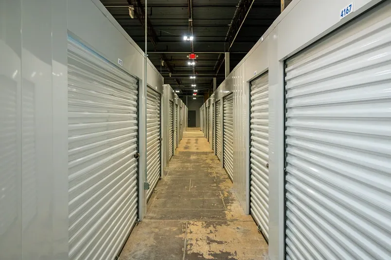 Indoor storage hallway with rows of units and roll-up doors under overhead lighting