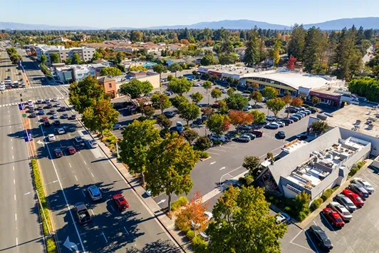 Aerial view of a suburban commercial district with tree-lined roads, retail centers, parking lots, and distant mountains.