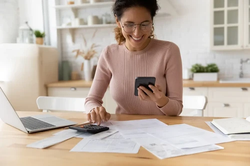A woman smiles while using her phone and documents to make a budget.