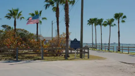 A flagpole with a flag flows in the wind beside palm trees outside of Boca Ciega Bay in Gulfport, FL.
