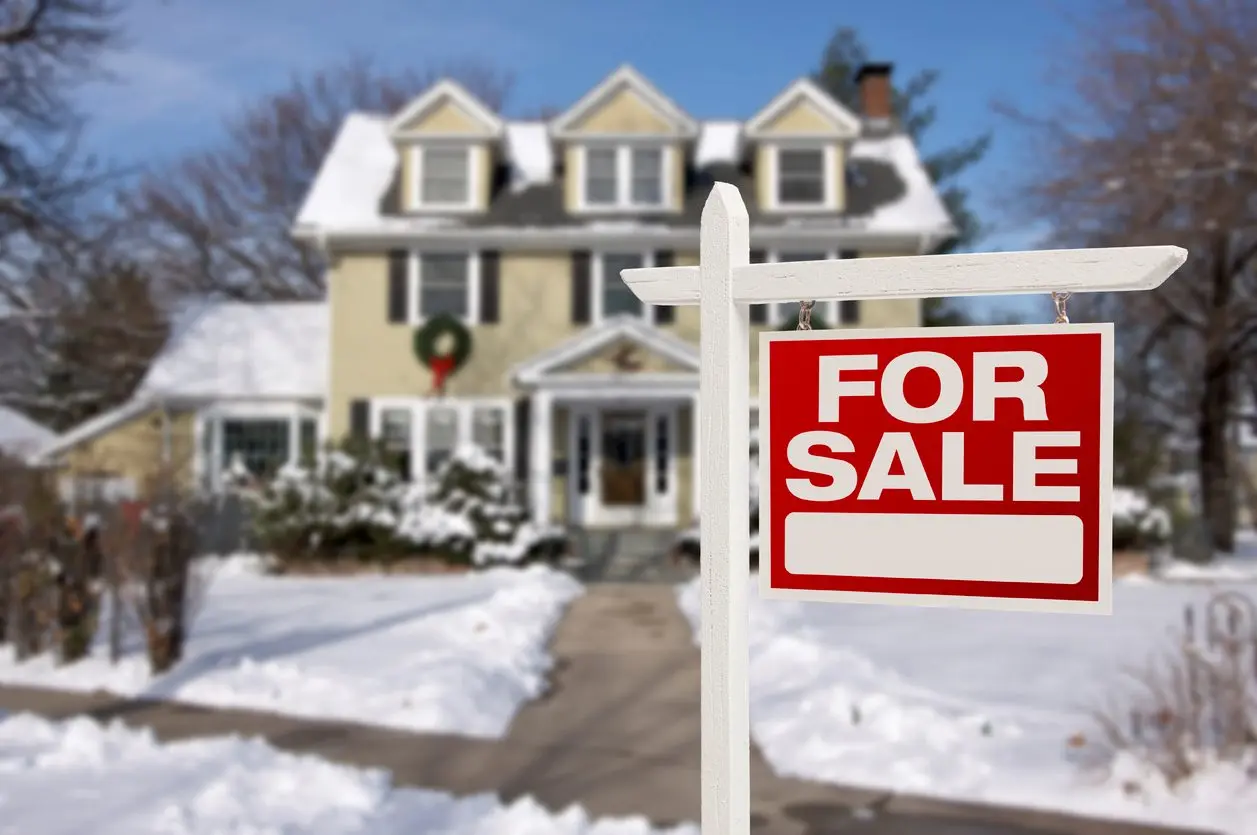 A home with a “for sale” sign surrounded by snow in the winter.
