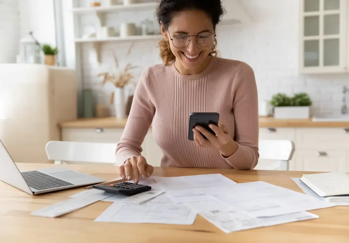 A woman smiles while using her phone and documents to make a budget.
