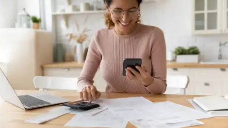 A woman smiles while using her phone and documents to make a budget.