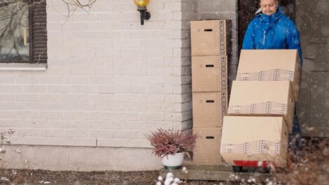 A man in a blue jacket wheels boxes on a cart out of his white brick home as it snows outside.