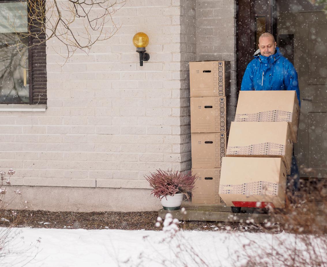 A man in a blue jacket wheels boxes on a cart out of his white brick home as it snows outside.